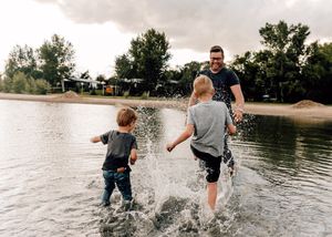 Father and two young boys splashing in a lake on a cloudy day.