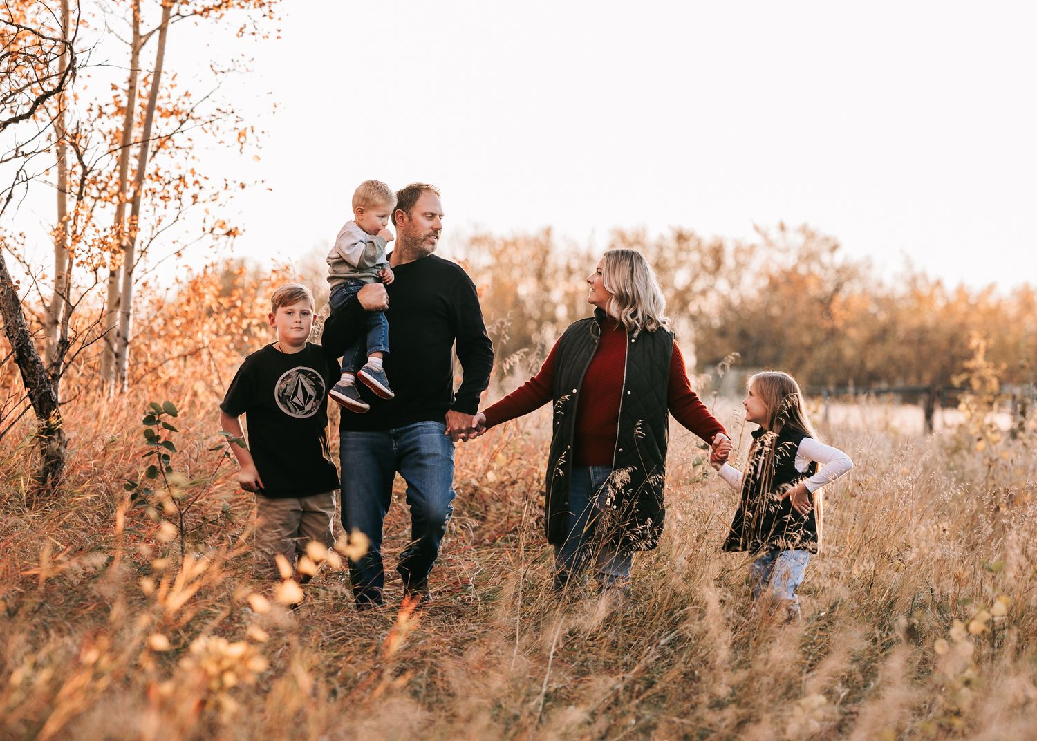 Family of five strolling through tall golden grass in autumn, holding hands at sunset.