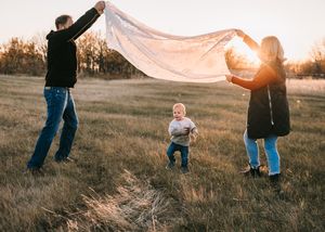 Parents holding a white blanket over their toddler in a golden sunset field, creating a magical moment.