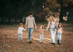 Family of six walking hand-in-hand through a sunlit autumn park, dressed in casual neutral tones.