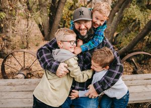Father laughing while hugging his three sons near a rustic fence in autumn.