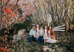 Family of five posing among colorful autumn foliage and stone wall outdoors.