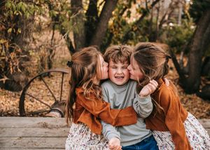 Two sisters joyfully kissing their brother on the cheeks outdoors in fall.