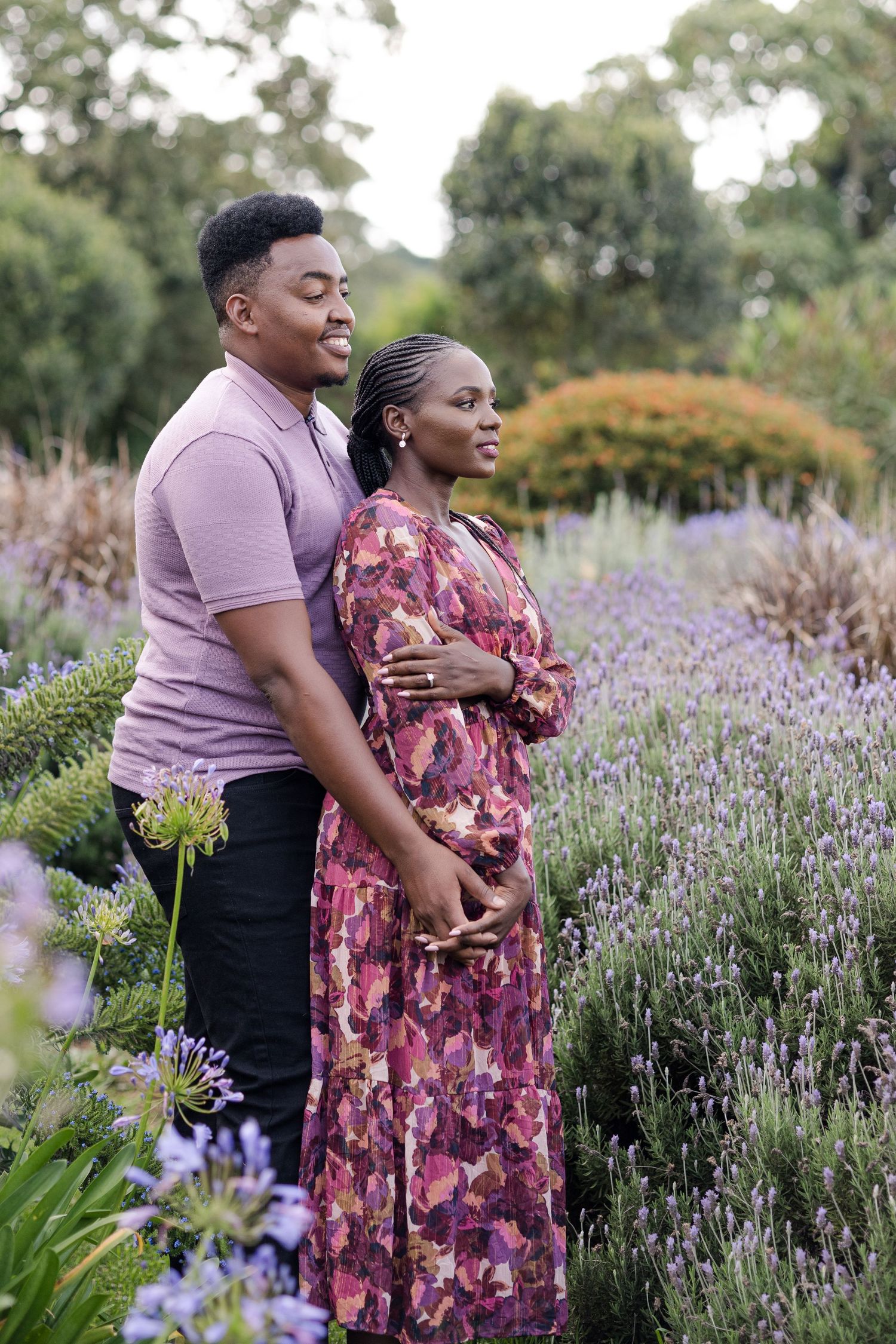Portrait of a couple in Naishola's lavender garden. Hugging from behind. Their right hands are holding. Looking off into the distance.