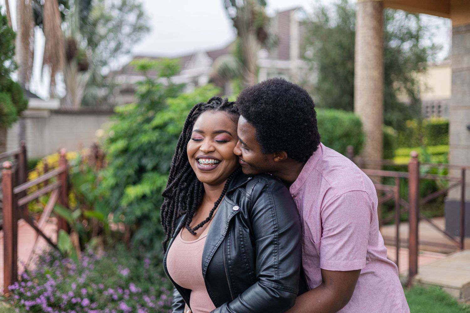 Portrait of a couple in a garden. Man is hugging her from behind, giving her a kiss on the cheek. She's smiling widely.