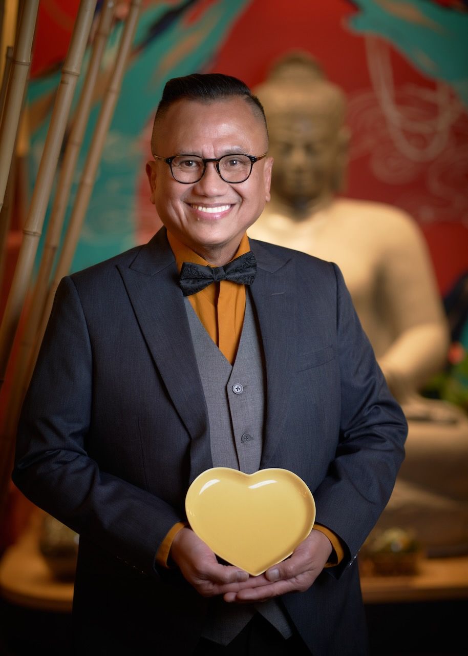 Professional portrait of someone in a dark suit and bow tie holding a golden heart-shaped object against a reddish background.