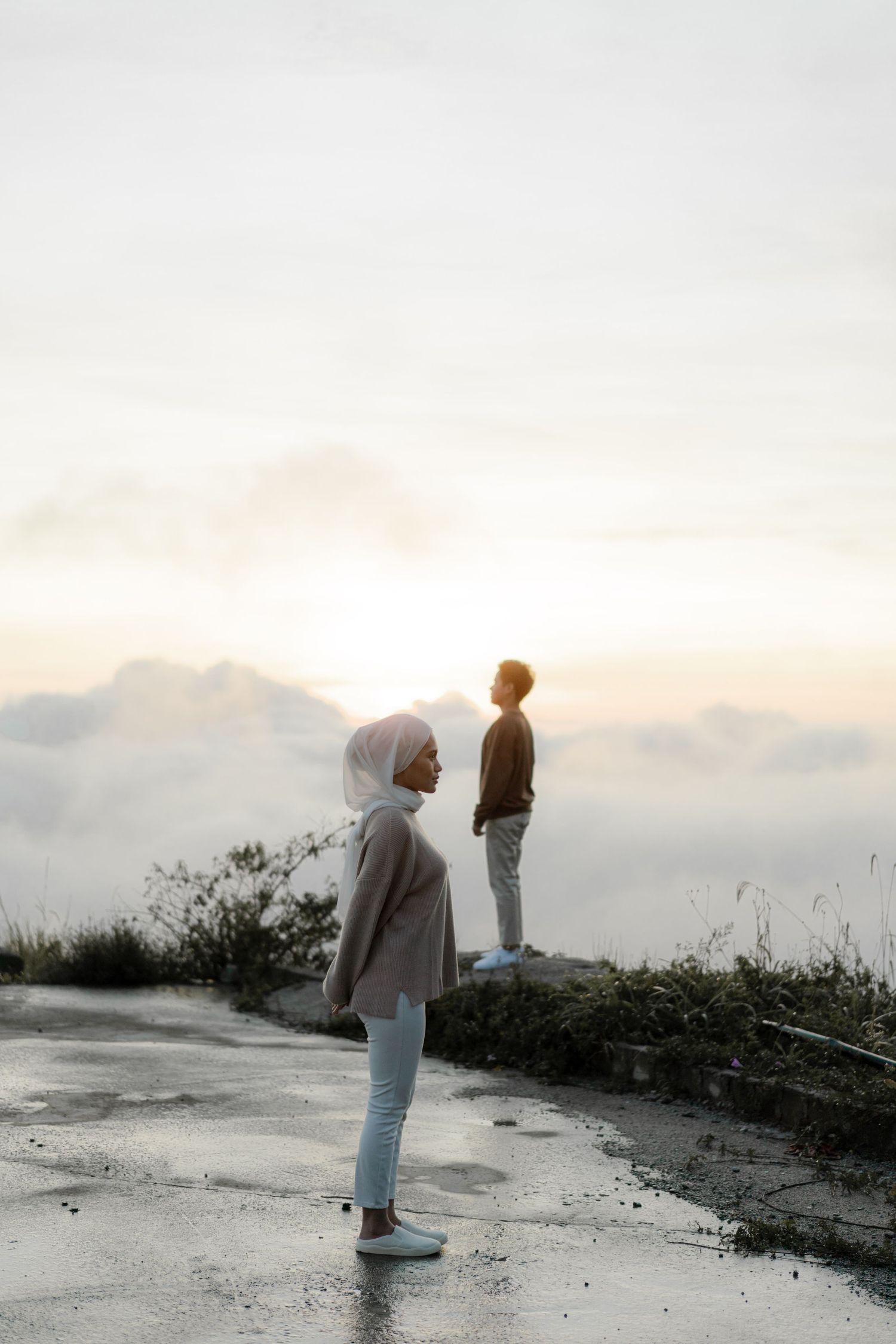 Two people stand on a misty mountain path at sunset looking at the horizon.