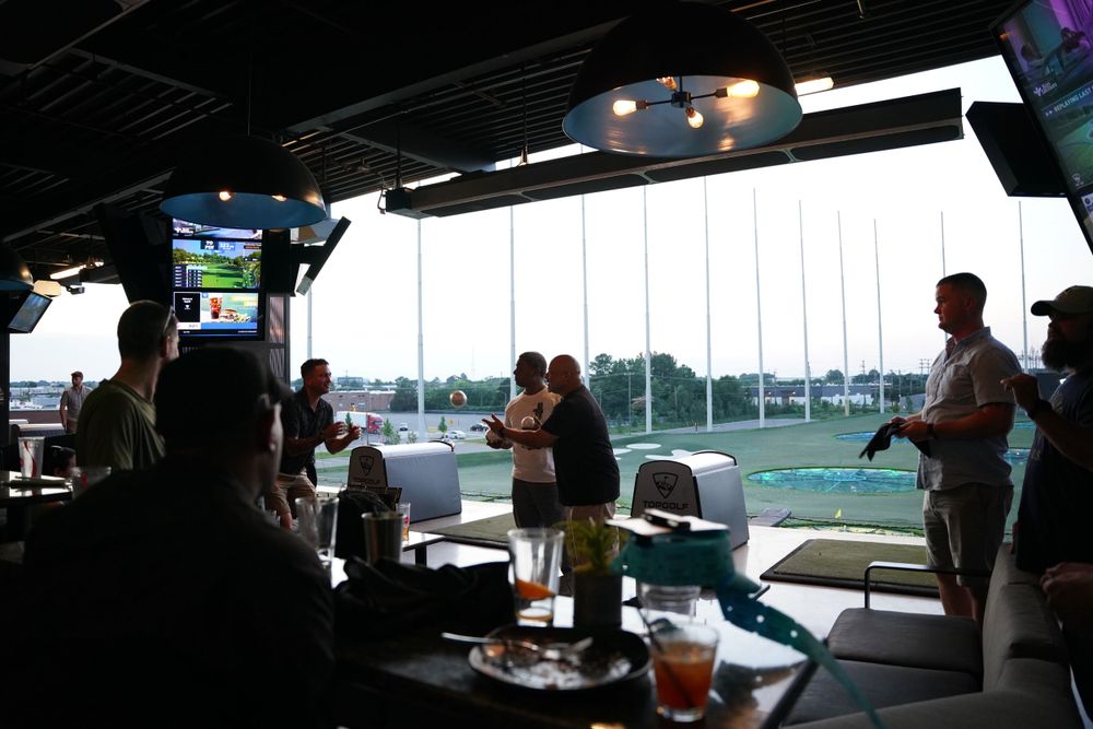 People gather in a golf driving range bay overlooking a course at dusk.