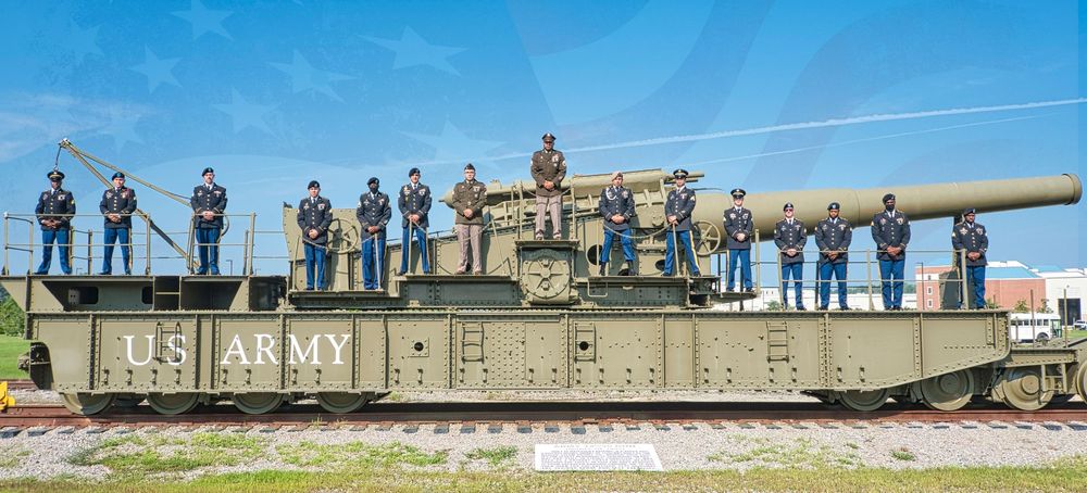 U.S. Army soldiers stand atop a historic railway artillery gun on a flatcar at an outdoor military display.