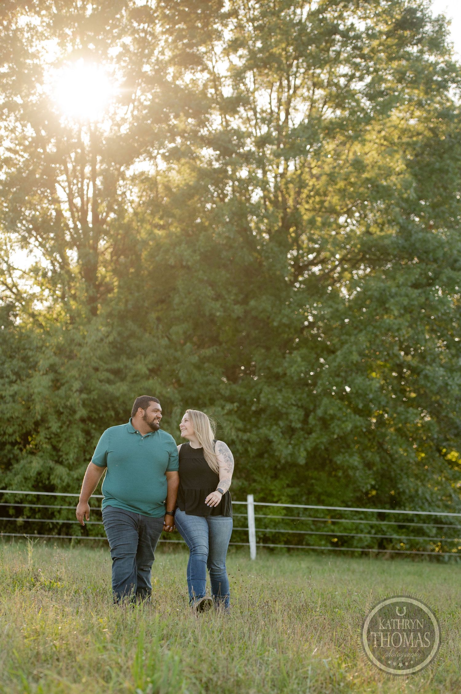 A couple walks together in a sunlit field surrounded by trees.