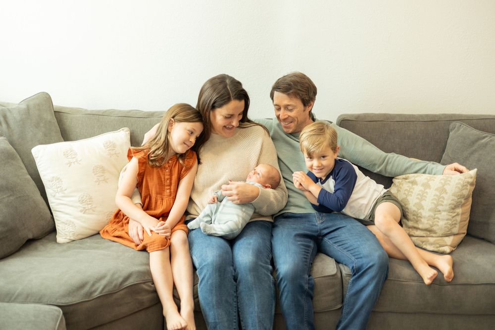 Family of five cuddled together on gray couch looking at newborn baby.