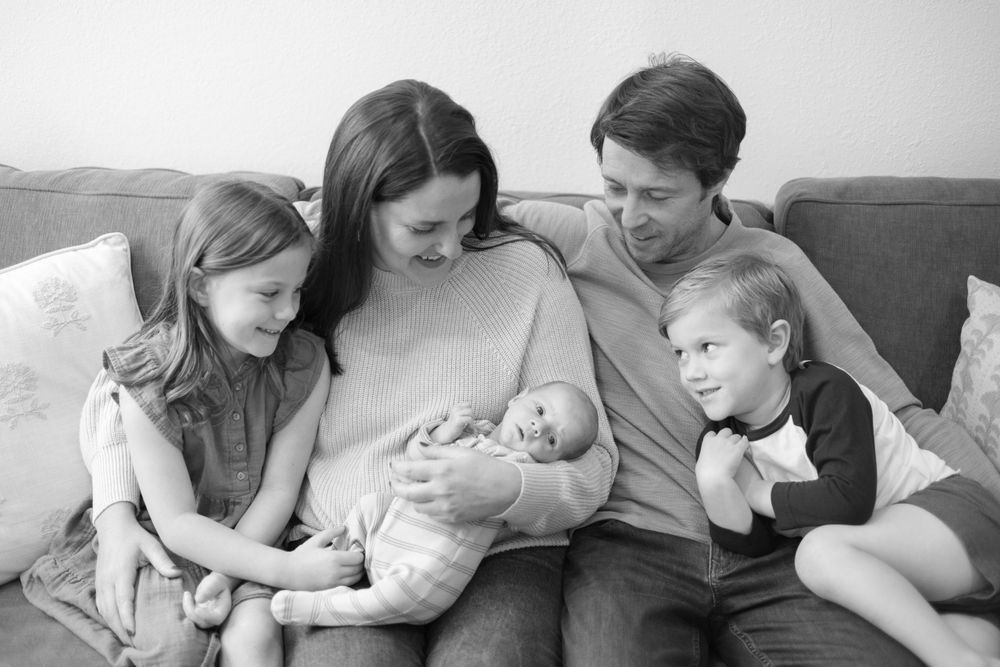 A family of five sits together on a gray couch viewing their newborn baby in an intimate black and white moment.