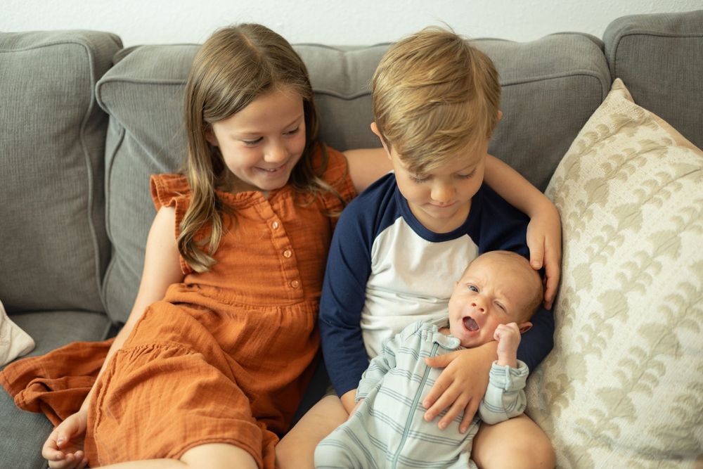 Three siblings sitting on a couch sharing a tender moment with their new baby sibling.
