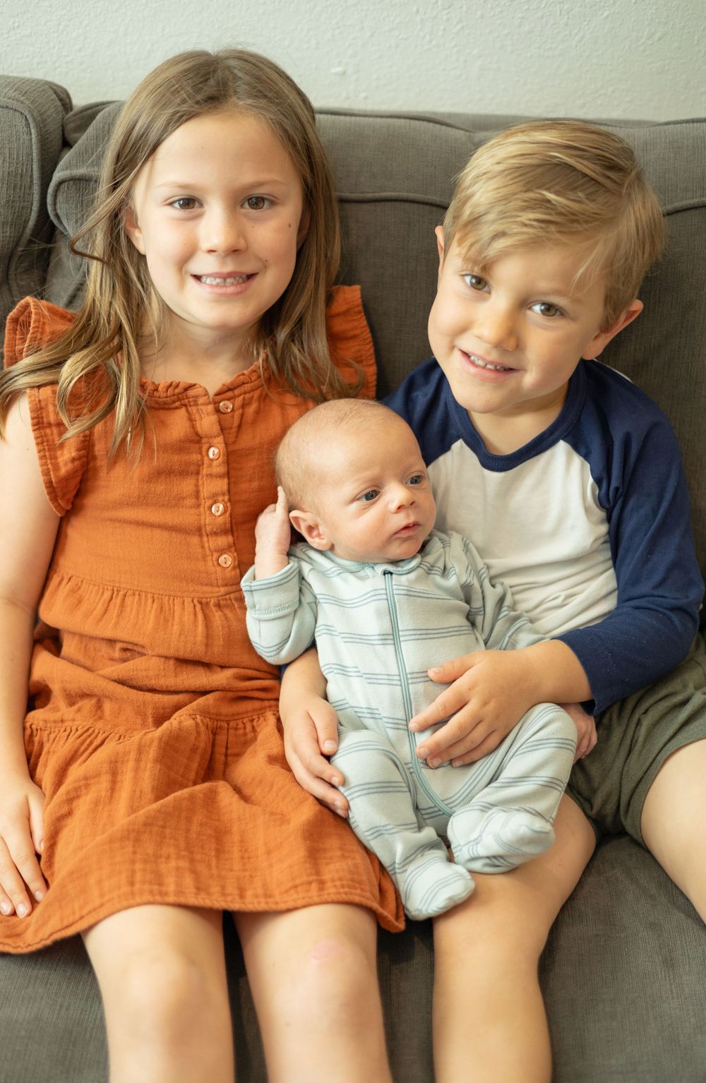 Children in orange and blue clothing pose with an infant sibling on a gray sofa.