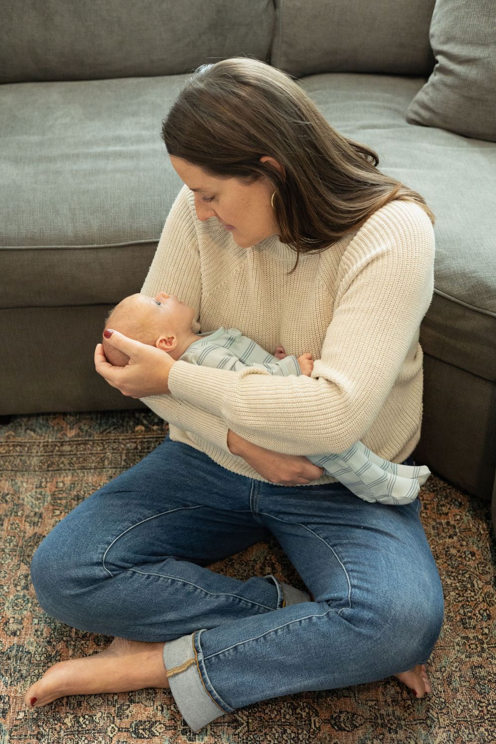 A woman in a cream sweater and jeans cuddles a baby while sitting on a patterned rug.