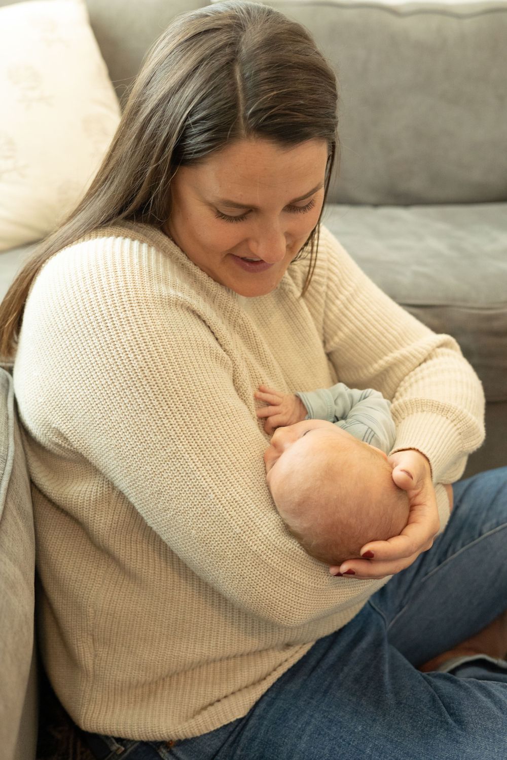 Woman in cream sweater holds and gazes lovingly at baby on gray couch.