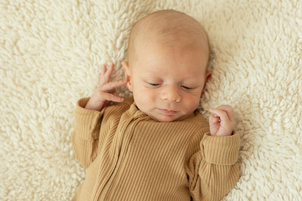 Sleeping newborn baby wearing brown onesie stretches on cream colored blanket.