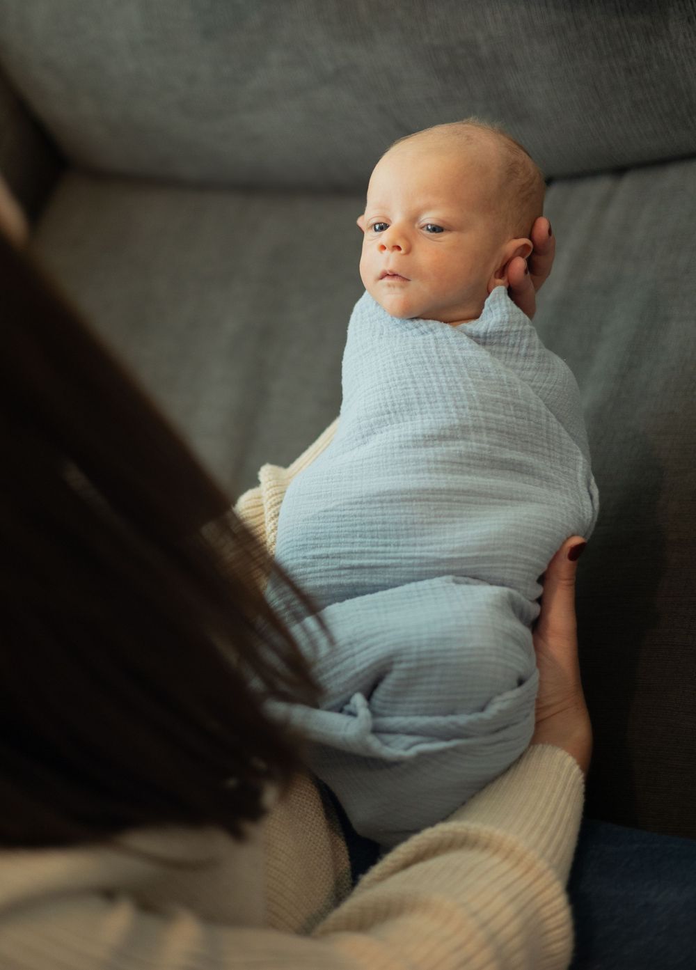 A small infant wrapped in soft blue muslin fabric resting on a gray couch.