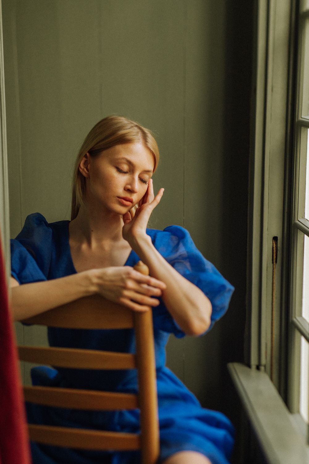 A figure in a bright blue dress sits pensively by a window in dramatic lighting.