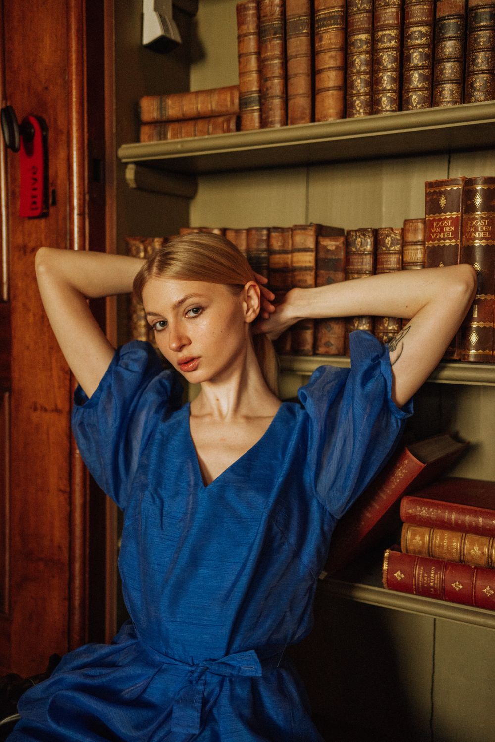 Figure in royal blue dress poses elegantly against vintage bookshelves in warm lighting.