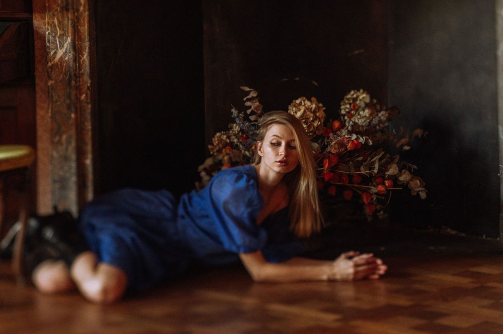 Artistic portrait of person in blue dress lying on wooden floor near floral arrangement.