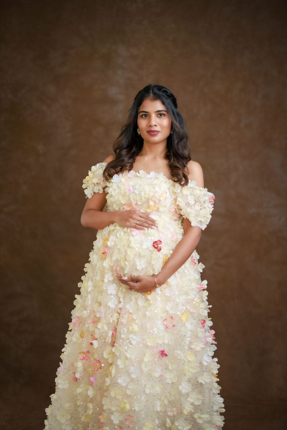 An ethereal maternity portrait in a cream floral applique gown against a brown backdrop.
