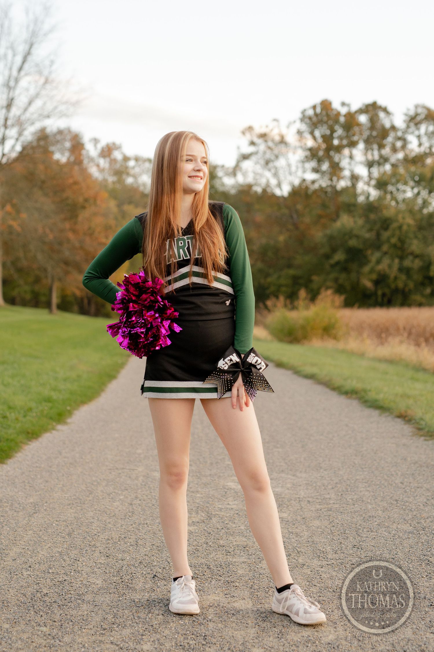 Cheerleader in green and black uniform poses with pink pom poms on a walking path surrounded by autumn trees.