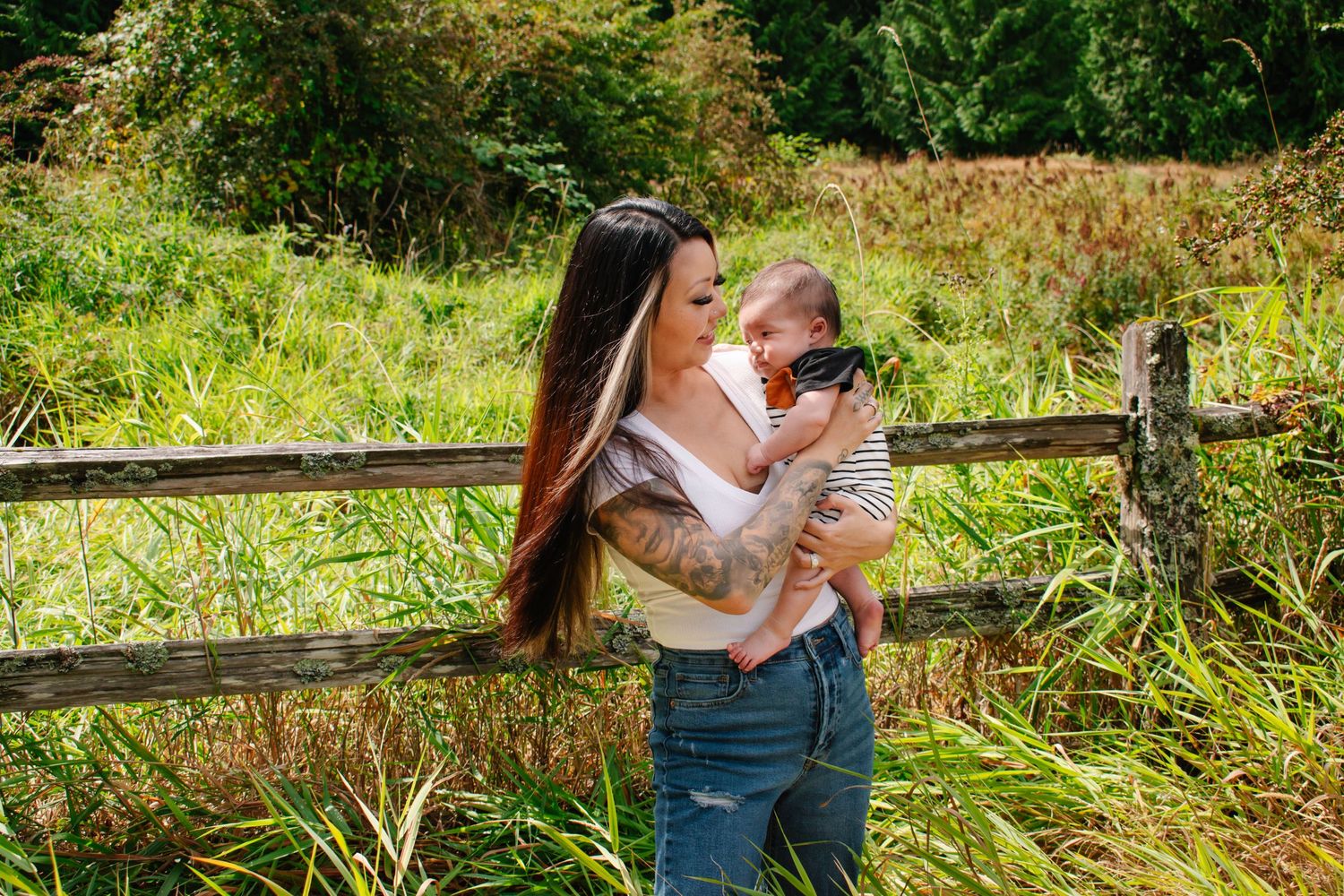 Seattle motherhood photography. A mother wearing jeans holds her son while standing near a wooden fence in a lush green field on a sunny day