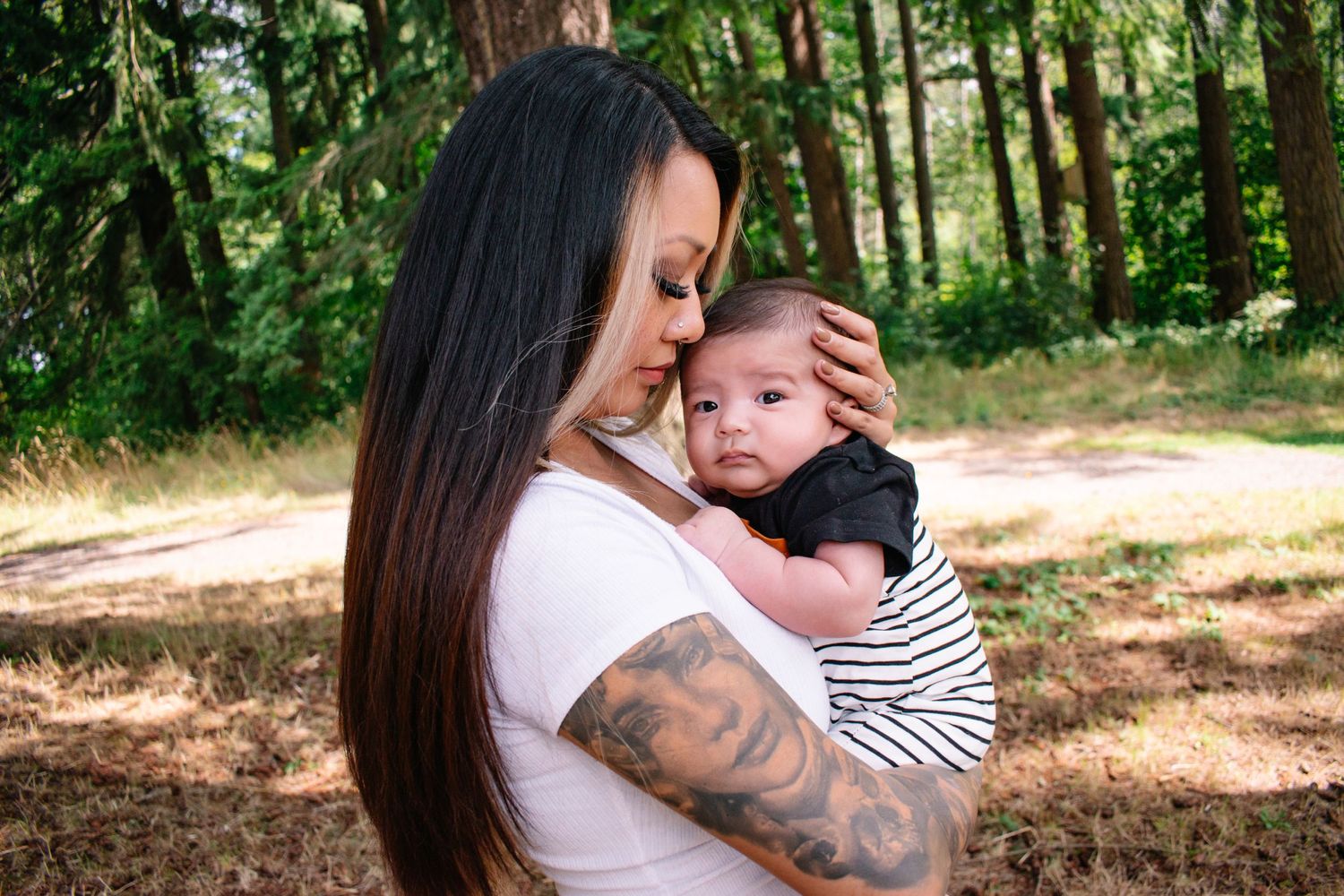 Seattle motherhood photo. Mother with long dark hair holds baby wearing striped shirt in sunny forest setting showing tender moment together