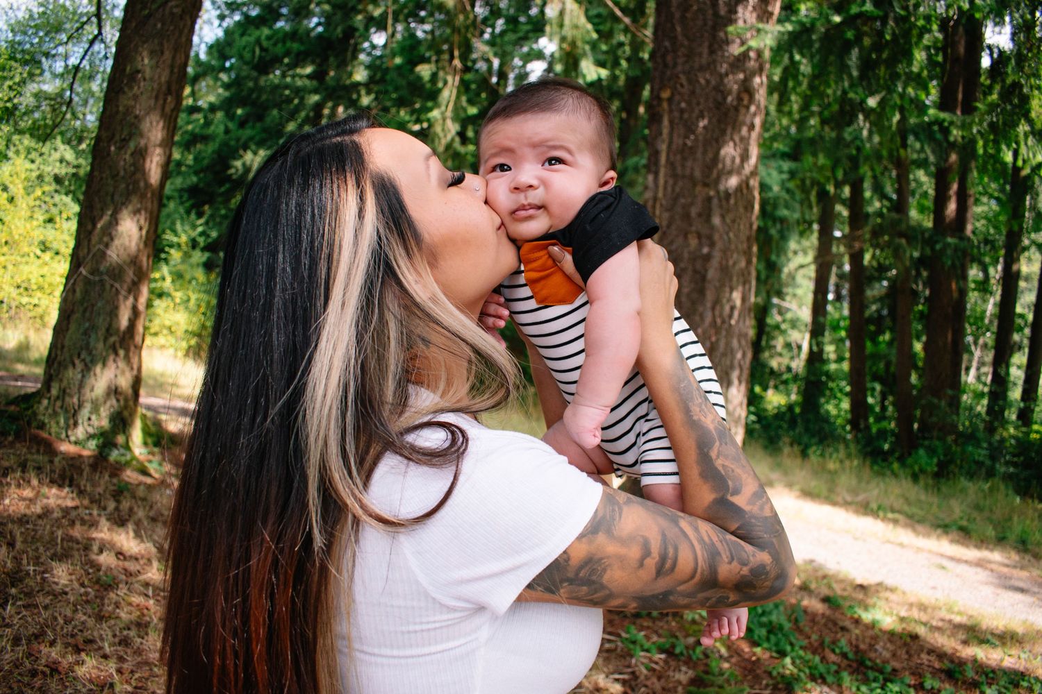 Seattle motherhood photography. A mother holding and kissing a smiling baby wearing a striped shirt in a sunny wooded setting.