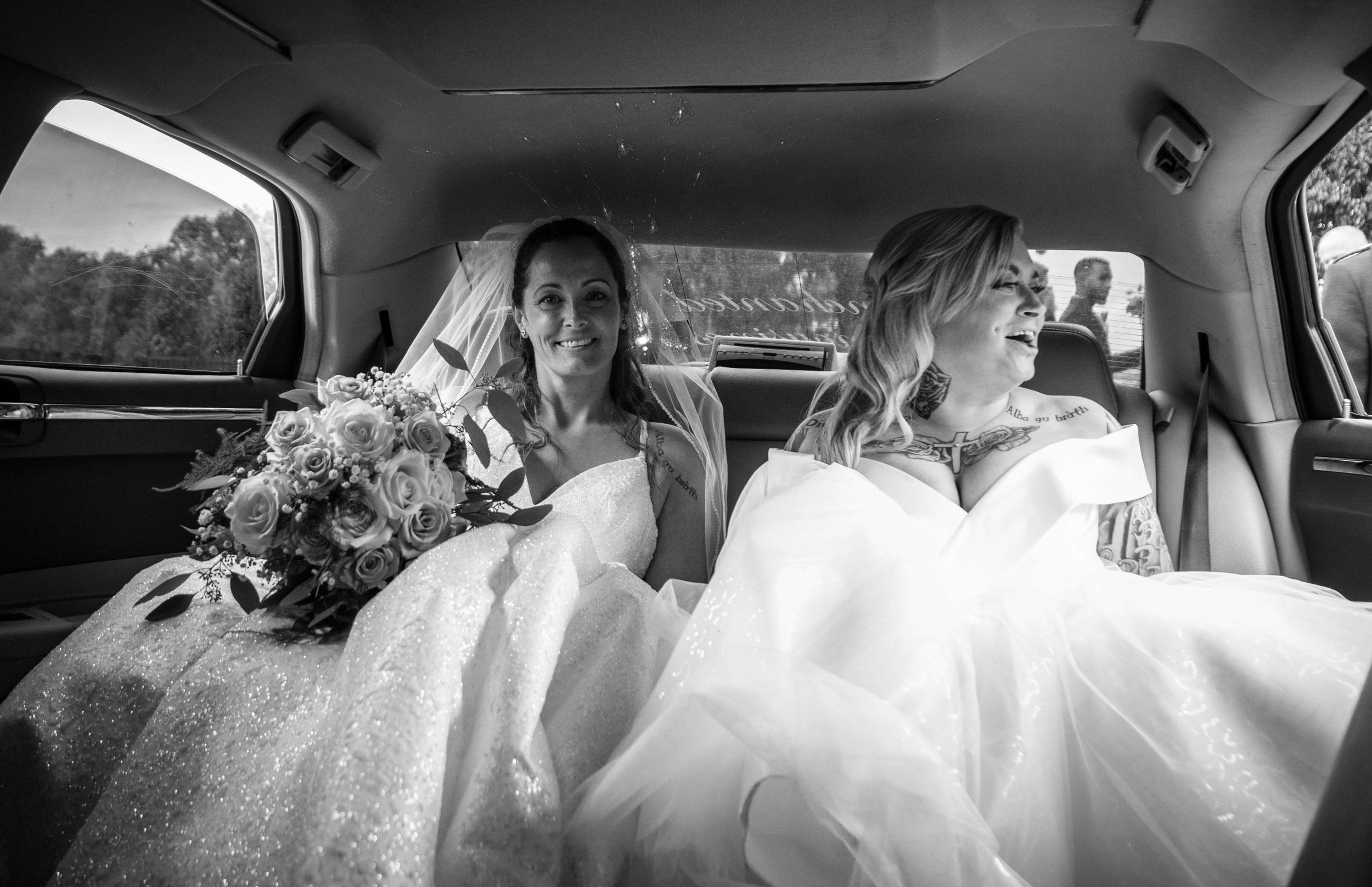 Two people in wedding attire sitting in the back of a car with bridal bouquet.