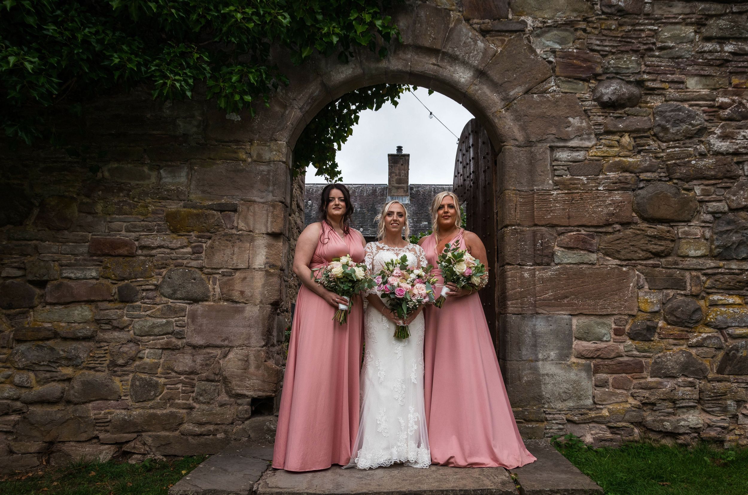 A bridal party poses under a rustic stone archway wearing pink bridesmaid dresses.