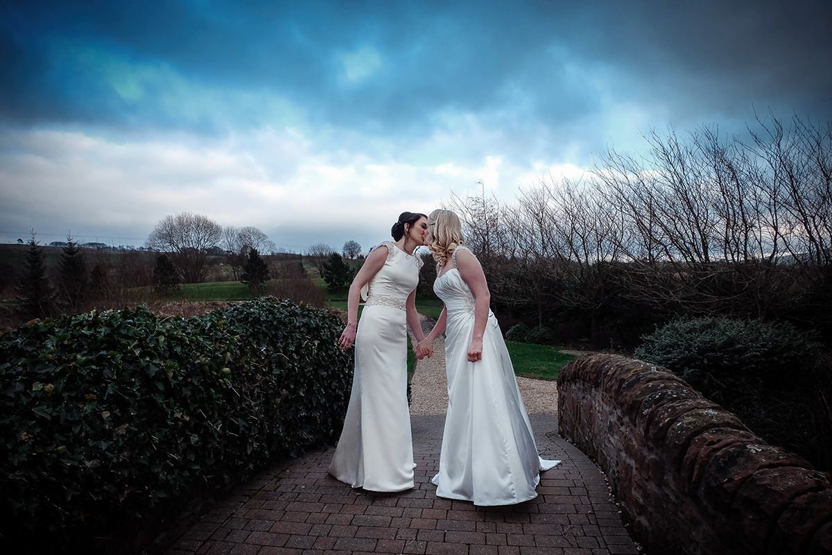 Two brides share a romantic moment on a stone path under dramatic stormy skies.
