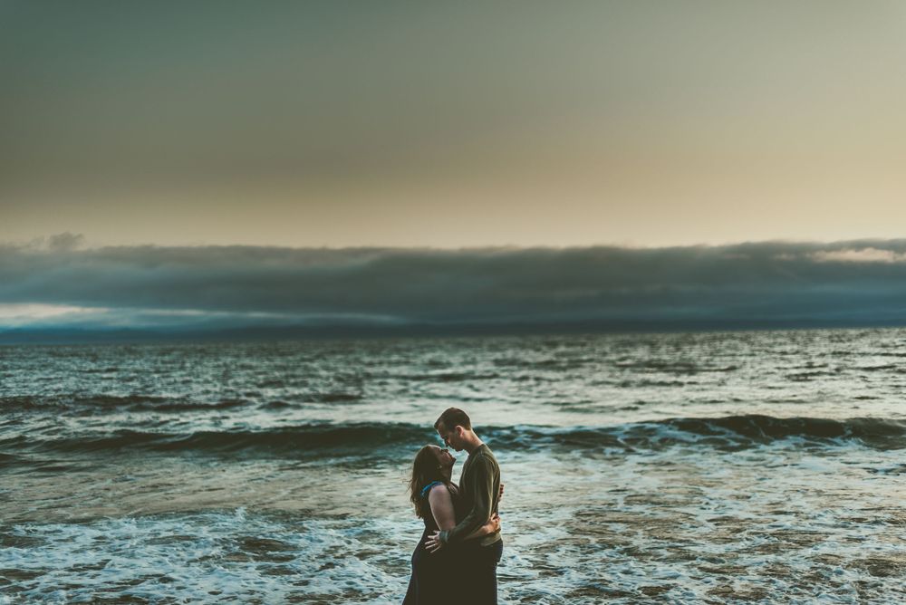 A silhouetted couple embraces on a moody beach with stormy clouds and crashing waves in the background Edmonton Engagement Photographer
