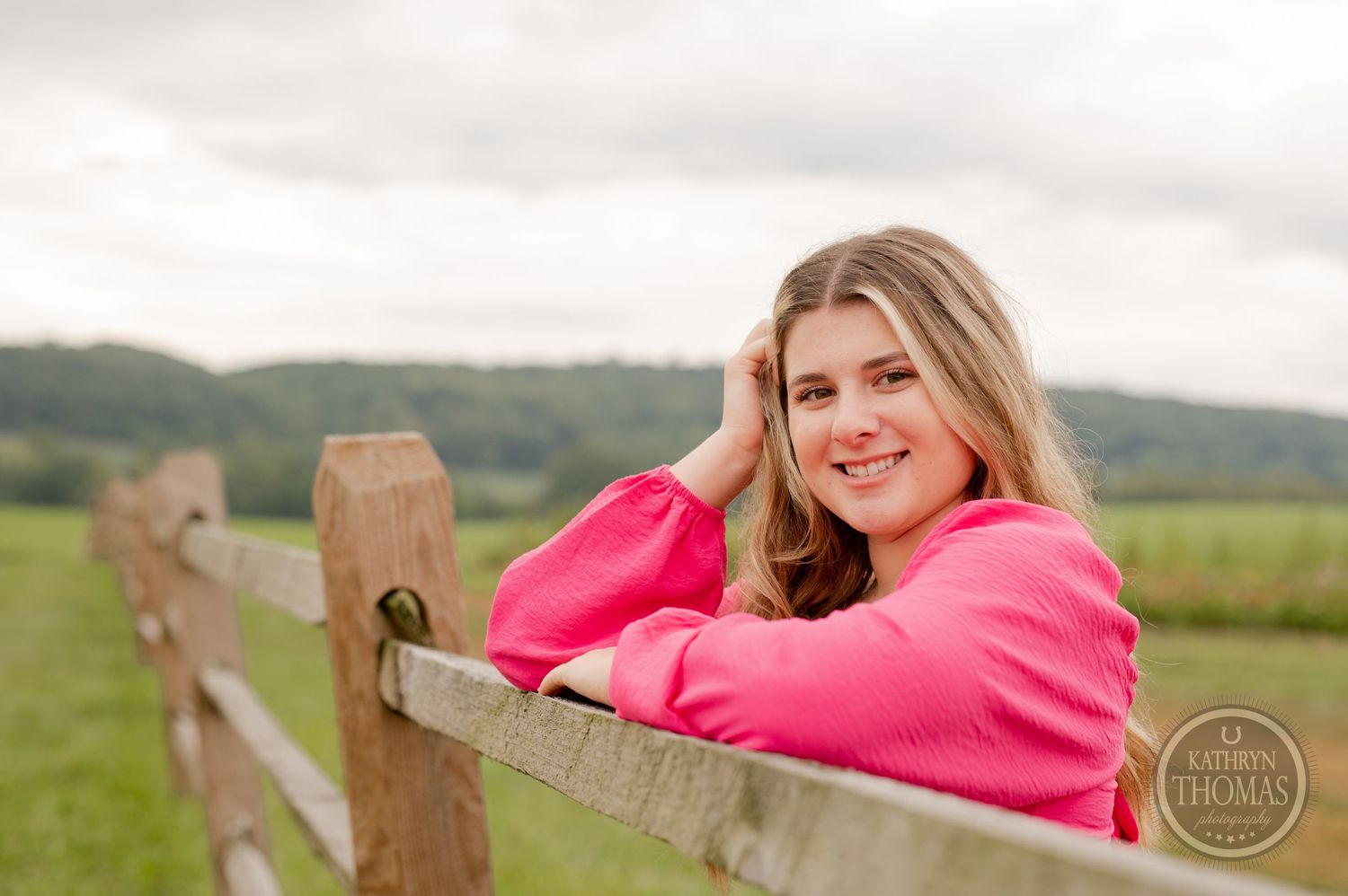 Outdoor portrait by wooden fence wearing bright pink sweater against cloudy rural landscape.