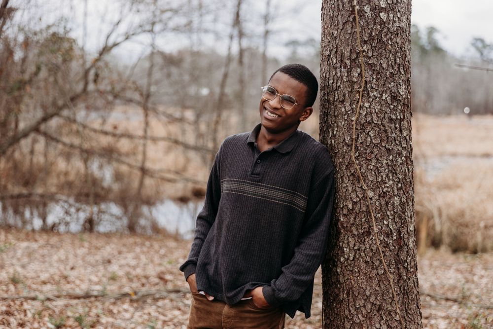 Person in casual attire poses next to tree trunk in natural outdoor setting.