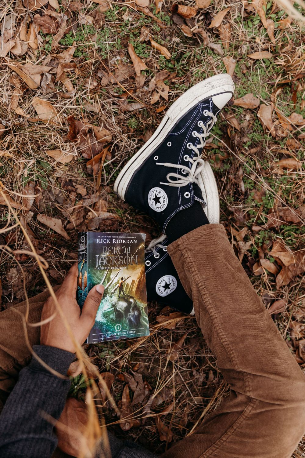 Converse high-top sneaker and Percy Jackson book placed on fallen autumn leaves.