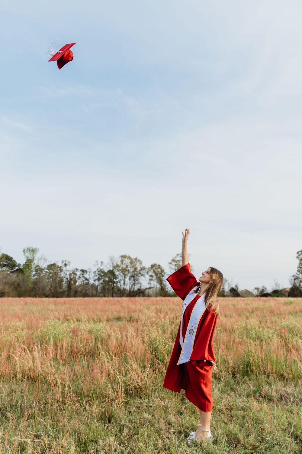 Graduate in red cap and gown tosses mortarboard into blue sky while standing in grassy field.