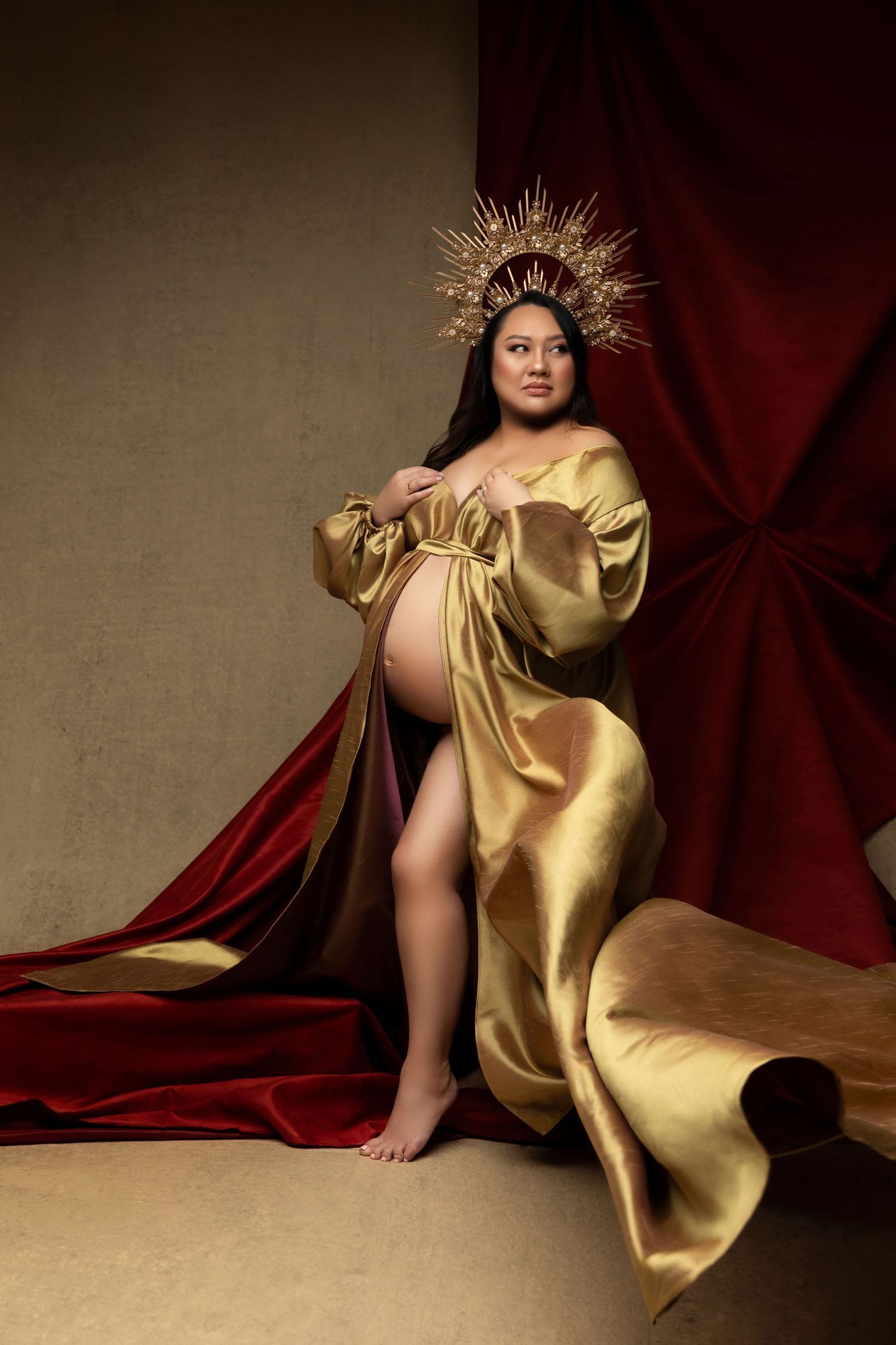 A maternity photoshoot featuring an elegant gold silk robe and ornate crown headpiece against deep red drapery.
