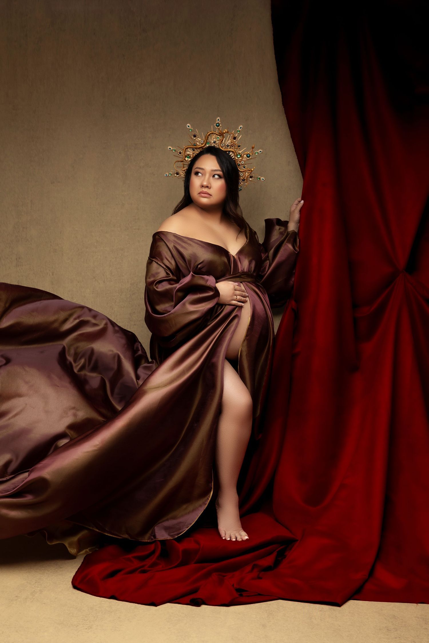 Dramatic portrait against red curtain backdrop features flowing brown silk gown and ornate golden crown headpiece.
