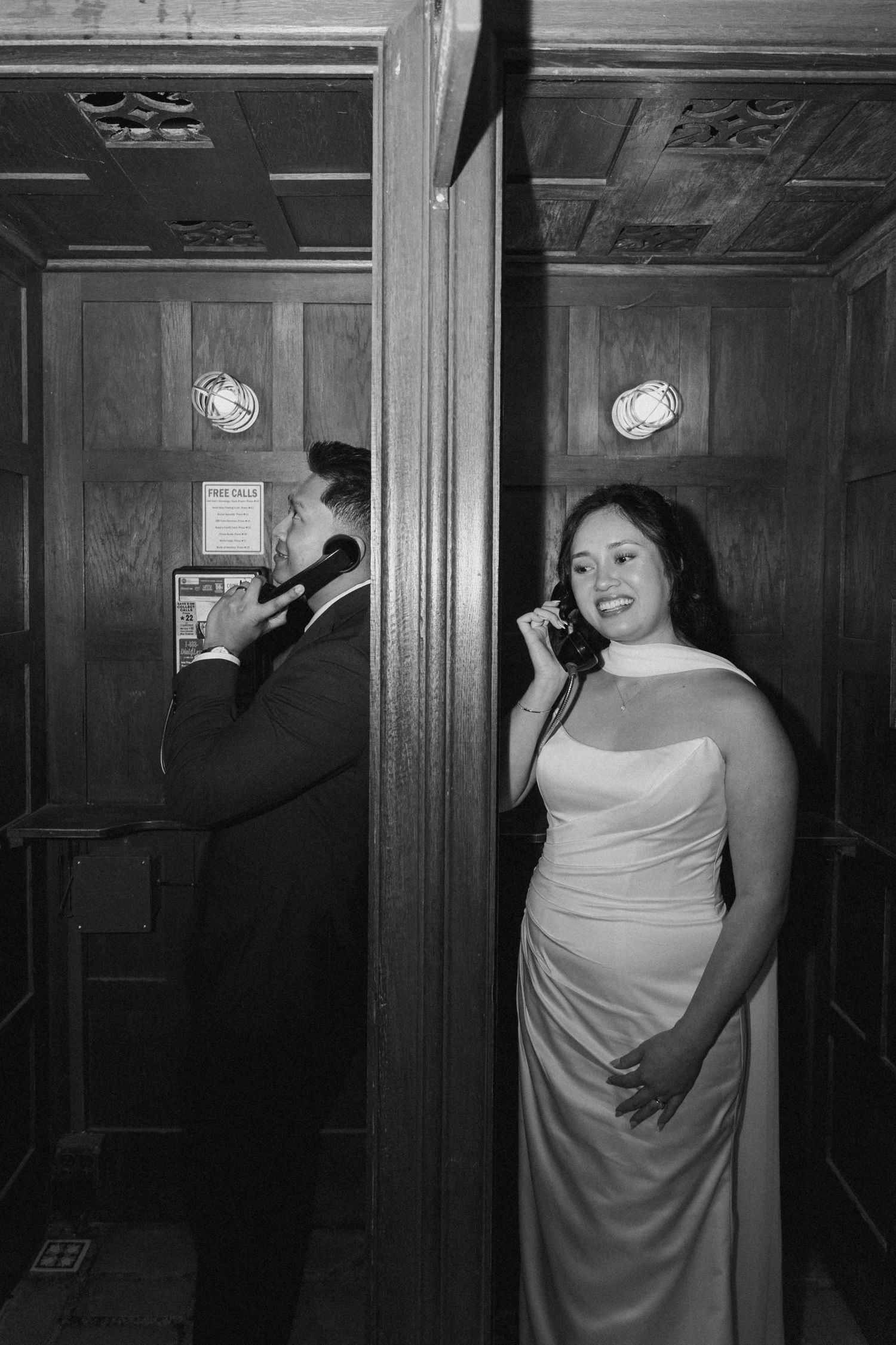 A black and white photo showing two people in adjacent phone booths, with one wearing a wedding dress.