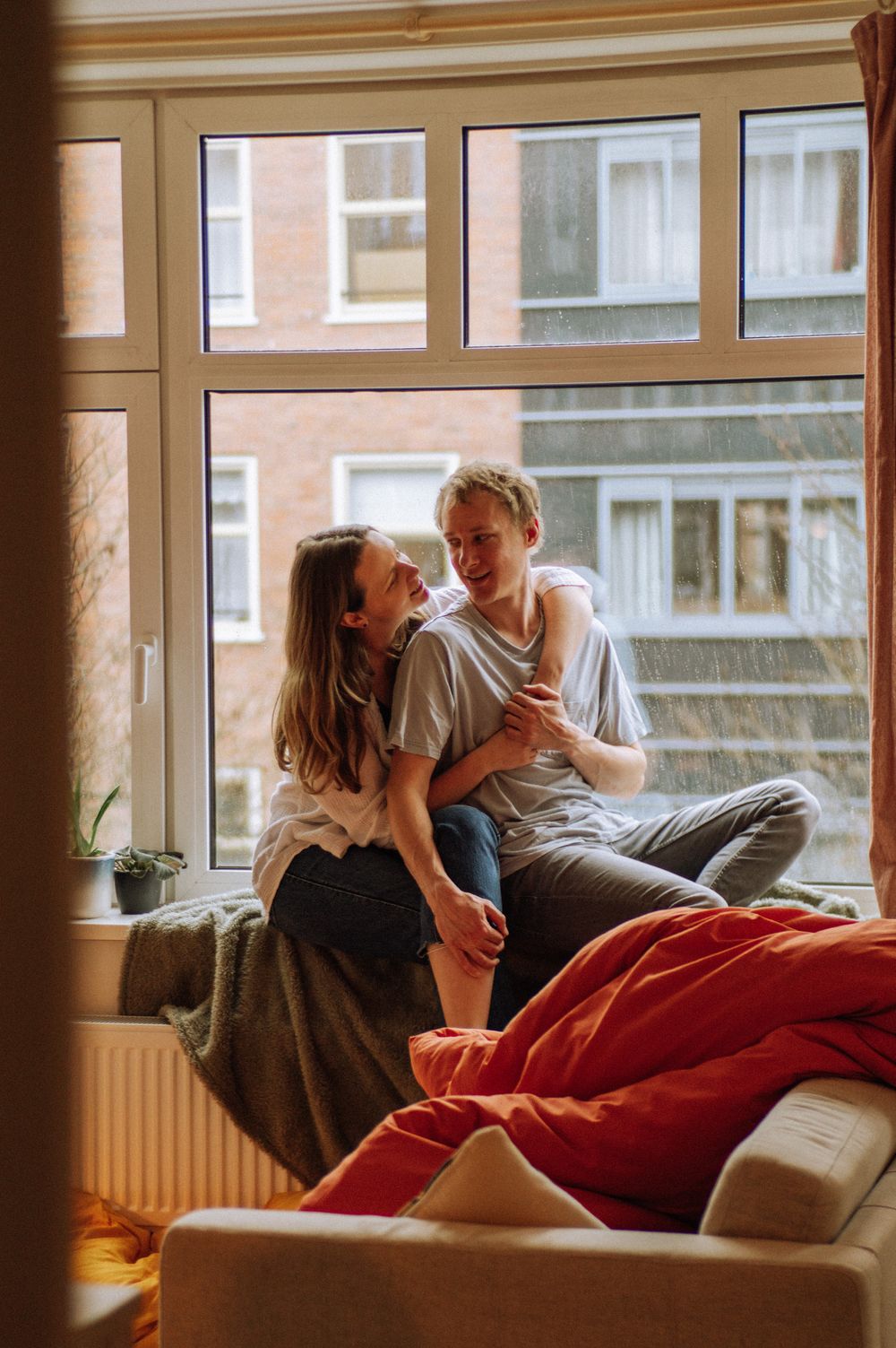 Couple sharing an intimate moment on a red couch by large windows.