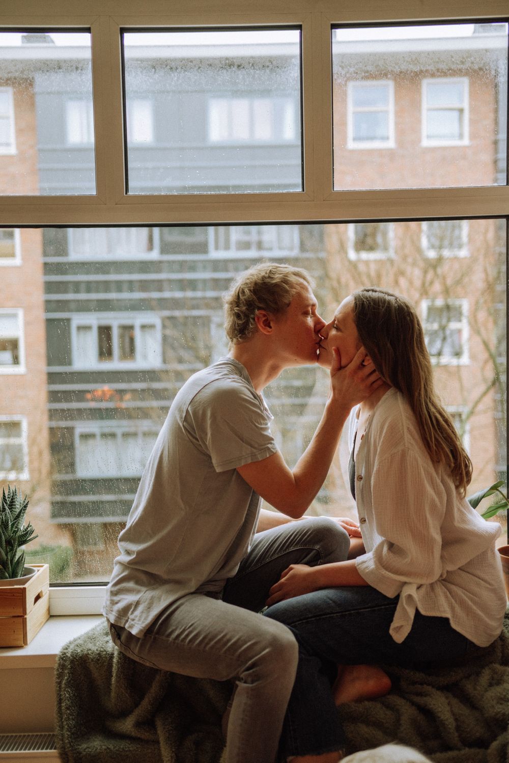 Couple sharing tender moment by large windows overlooking brick buildings.