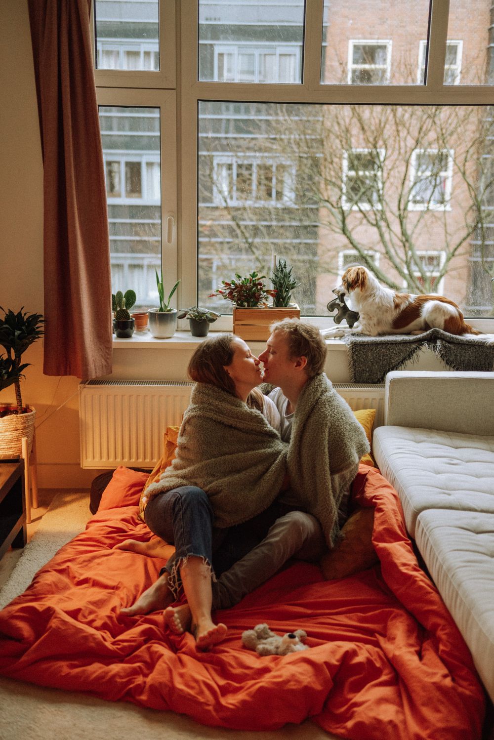 Intimate scene on red bedding with couple and dog relaxing by window.
