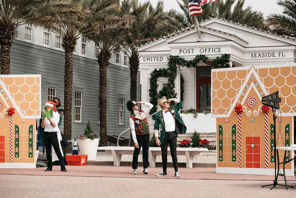 Performers dance and entertain in front of a decorated post office building with gingerbread house decorations.