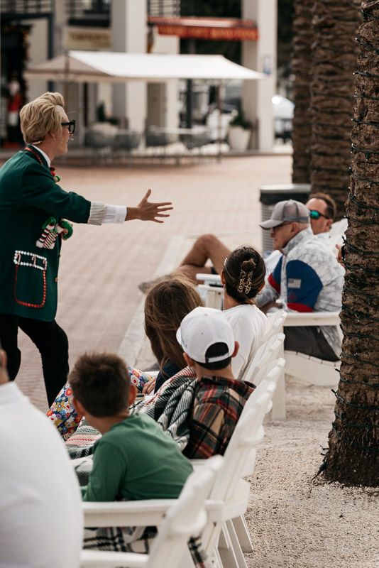 Performer in green jacket gestures enthusiastically to seated audience members during outdoor show.