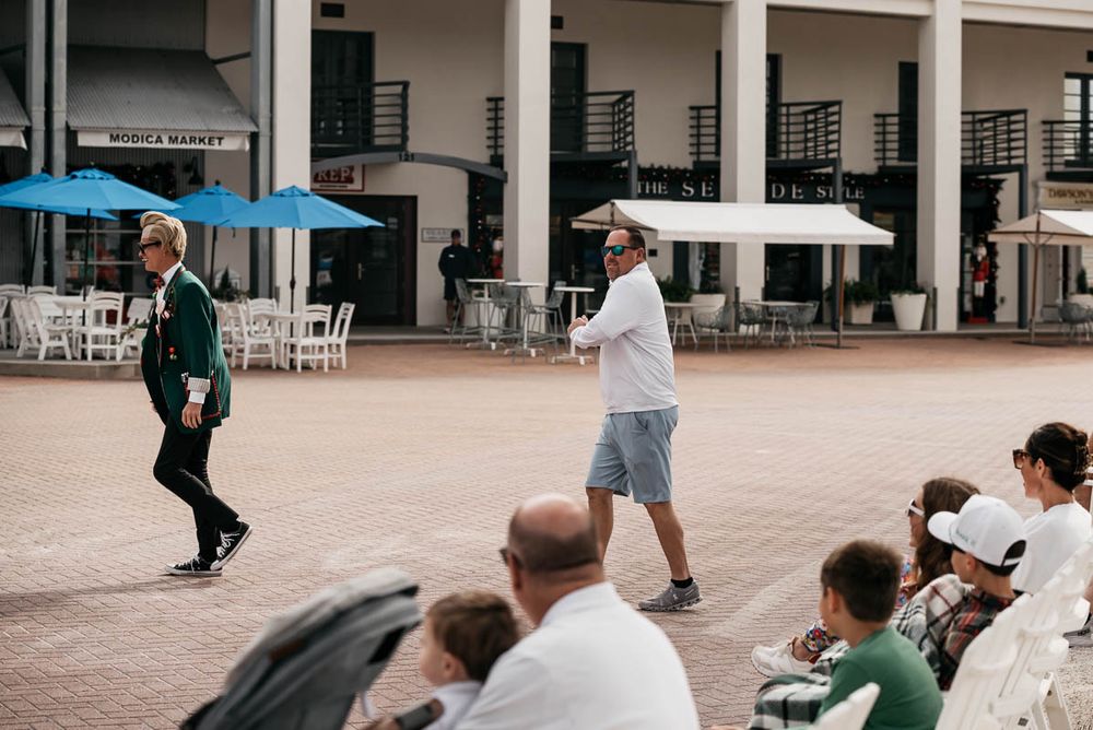 Performers entertain crowd in outdoor plaza with umbrellas and columns visible in background.