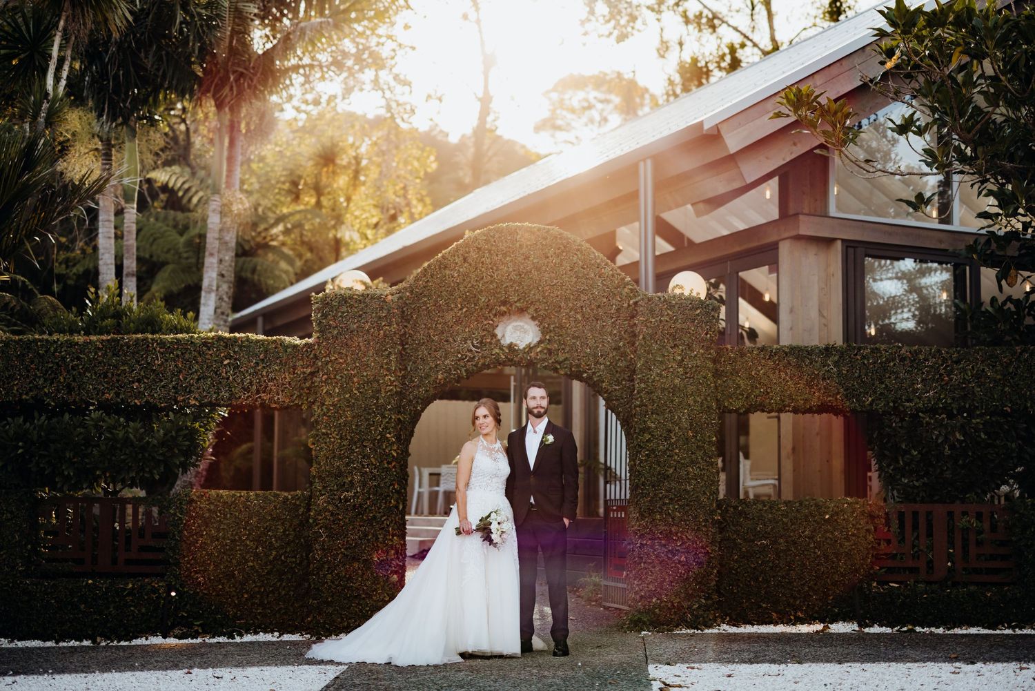 A wedding couple poses in front of a rustic building during golden hour with sunlight streaming through trees.