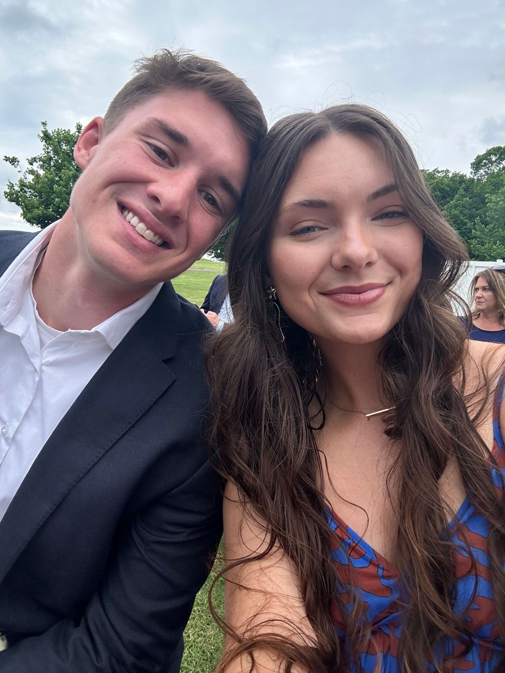 A young couple in formal attire smiles at the camera while taking a selfie outdoors on a cloudy day.