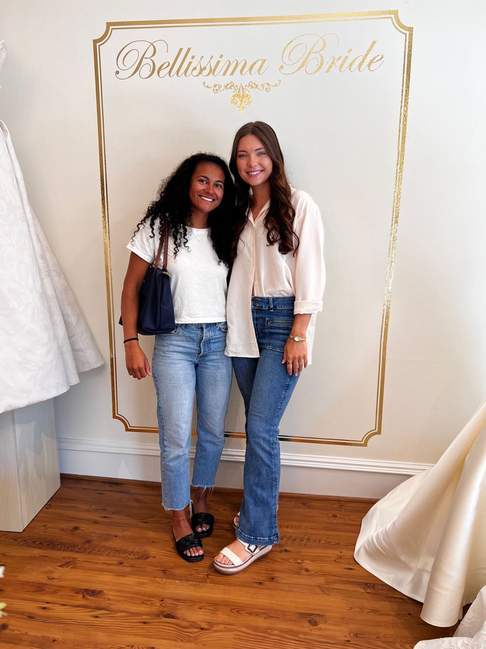 Two women standing in front of a Bellissima Bride backdrop wearing casual denim outfits.